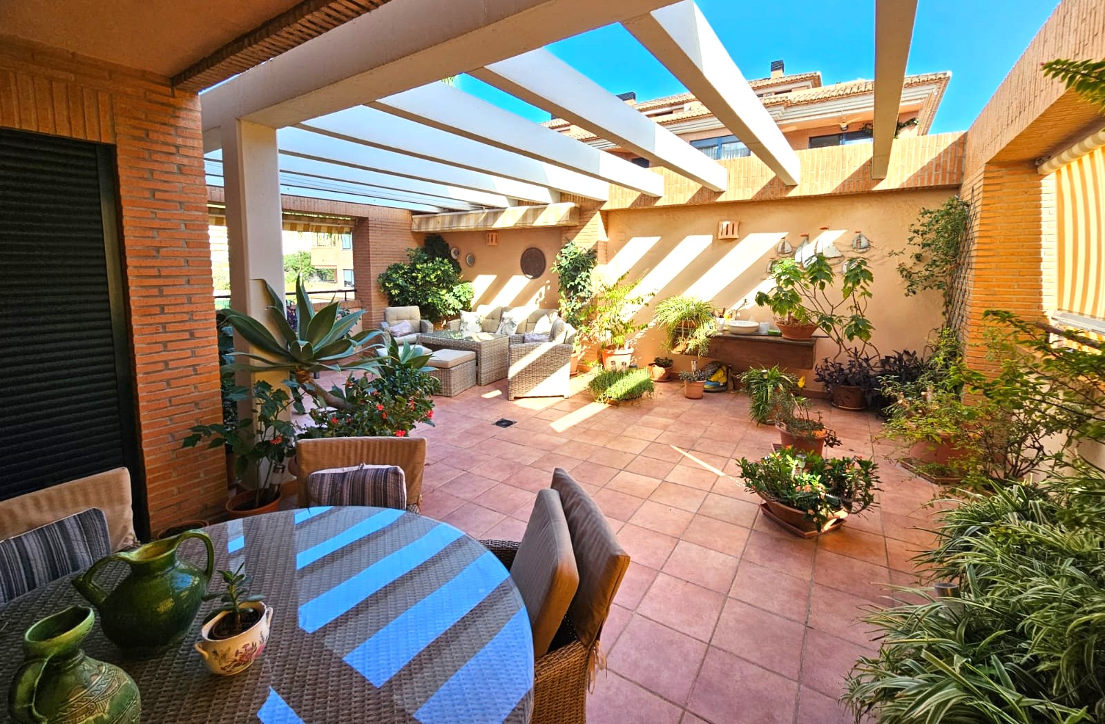 Spacious Mediterranean terrace featuring terracotta flooring, a white pergola, and integrated lounge and dining areas surrounded by lush potted plants.
