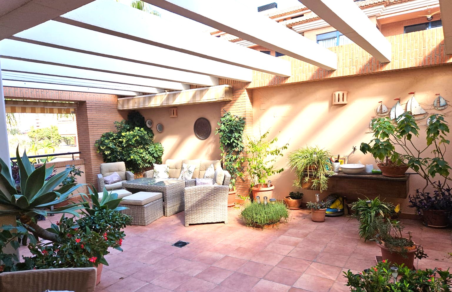 Spacious private terrace with terracotta flooring and white pergola beams. Features a wicker lounge set, integrated brickwork, and diverse Mediterranean potted plants.