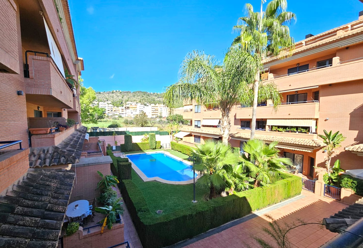 View from the balcony overlooking the communal swimming pool, manicured lawns, and palm trees, surrounded by brick-faced buildings with mountain views.