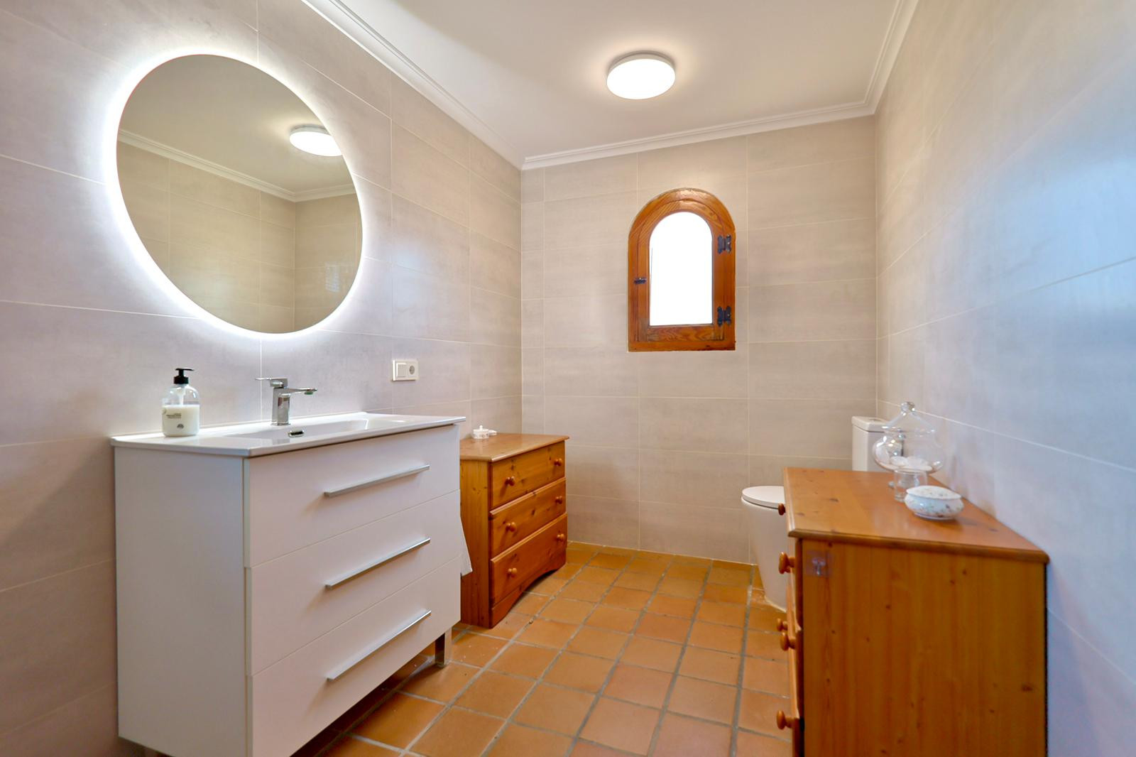Bathroom featuring a white vanity with an illuminated circular mirror and terracotta tile flooring. Includes a traditional arched wooden window and pine storage units.