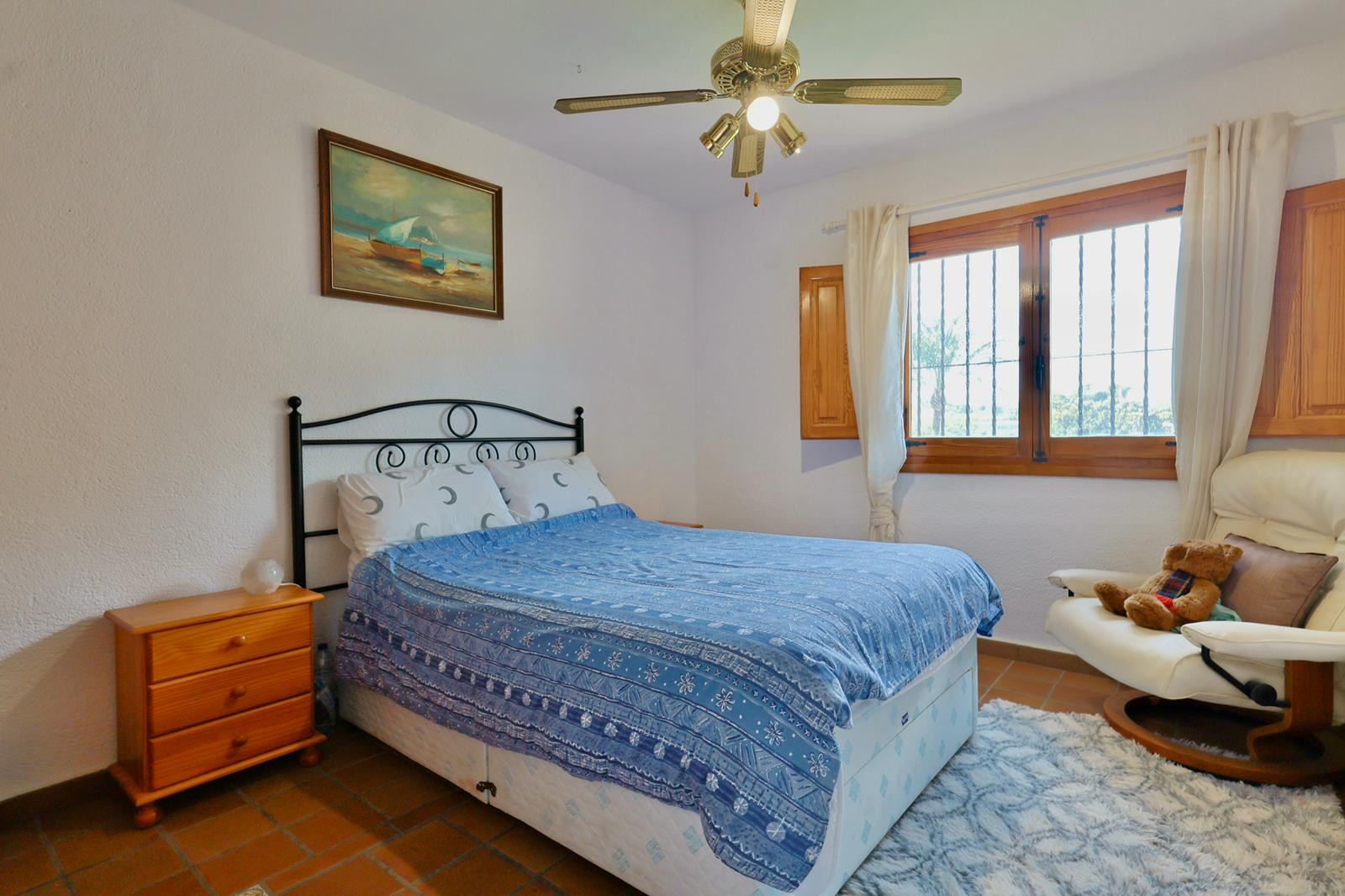 Bedroom featuring a wrought iron headboard, terracotta tile flooring, and a wooden window with shutters. Includes a ceiling fan and natural lighting.