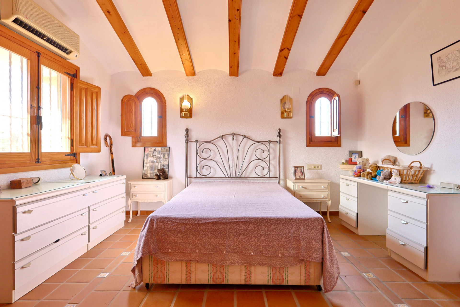 Bedroom featuring exposed wooden ceiling beams, terracotta tile floors, and arched windows with wooden shutters. Includes a wrought iron bed and split AC unit.