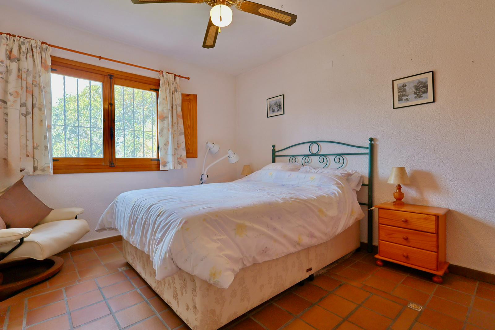 Bedroom featuring terracotta tile flooring, a wooden window frame with security bars, a green metal headboard, and a ceiling fan. Includes a matching wooden bedside table.