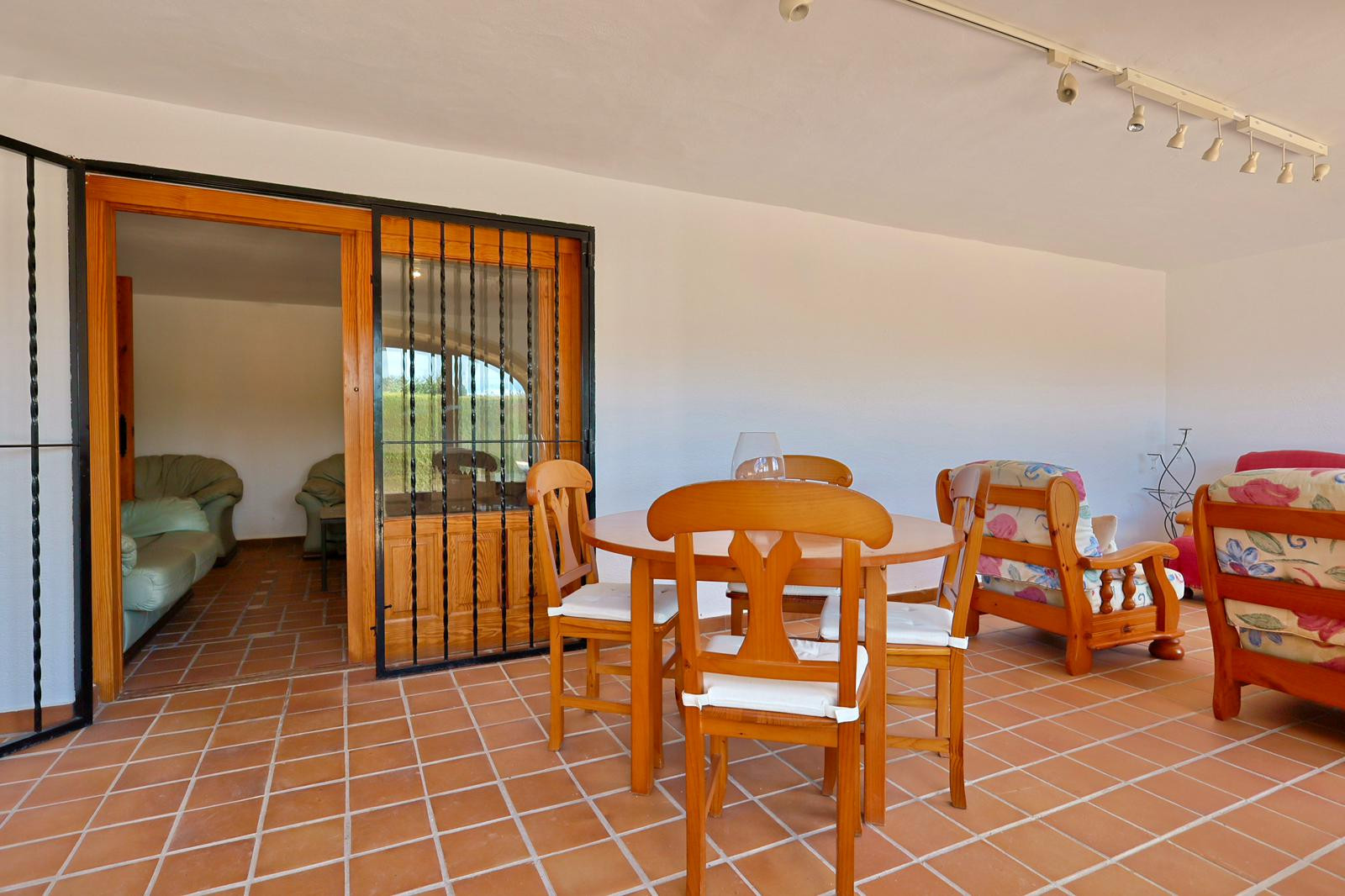 Covered terrace featuring terracotta tile flooring, a wooden dining set, and floral armchairs. A sliding glass door with wrought iron bars leads to the interior living area.