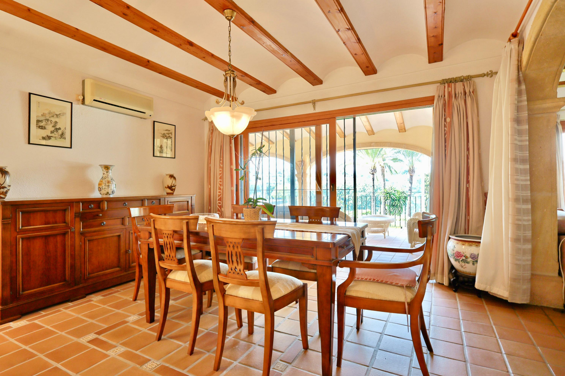 Dining room with exposed wooden beams, terracotta flooring, and a large sideboard. Sliding glass doors lead to a covered terrace with views of palm trees.
