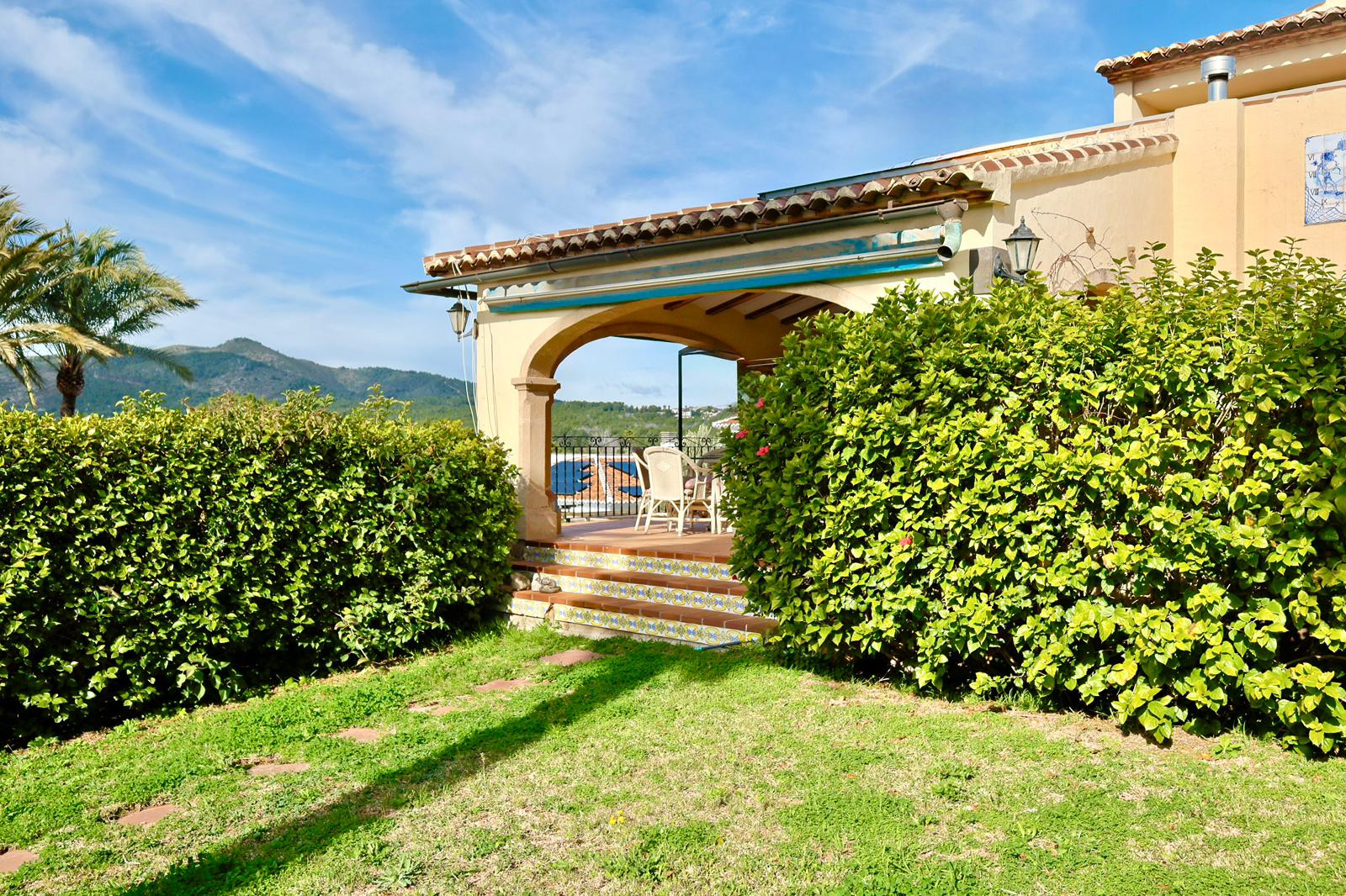 Exterior view of a covered terrace with a traditional stone arch, terracotta roof, and decorative tiled steps leading to a lawn with mountain views.
