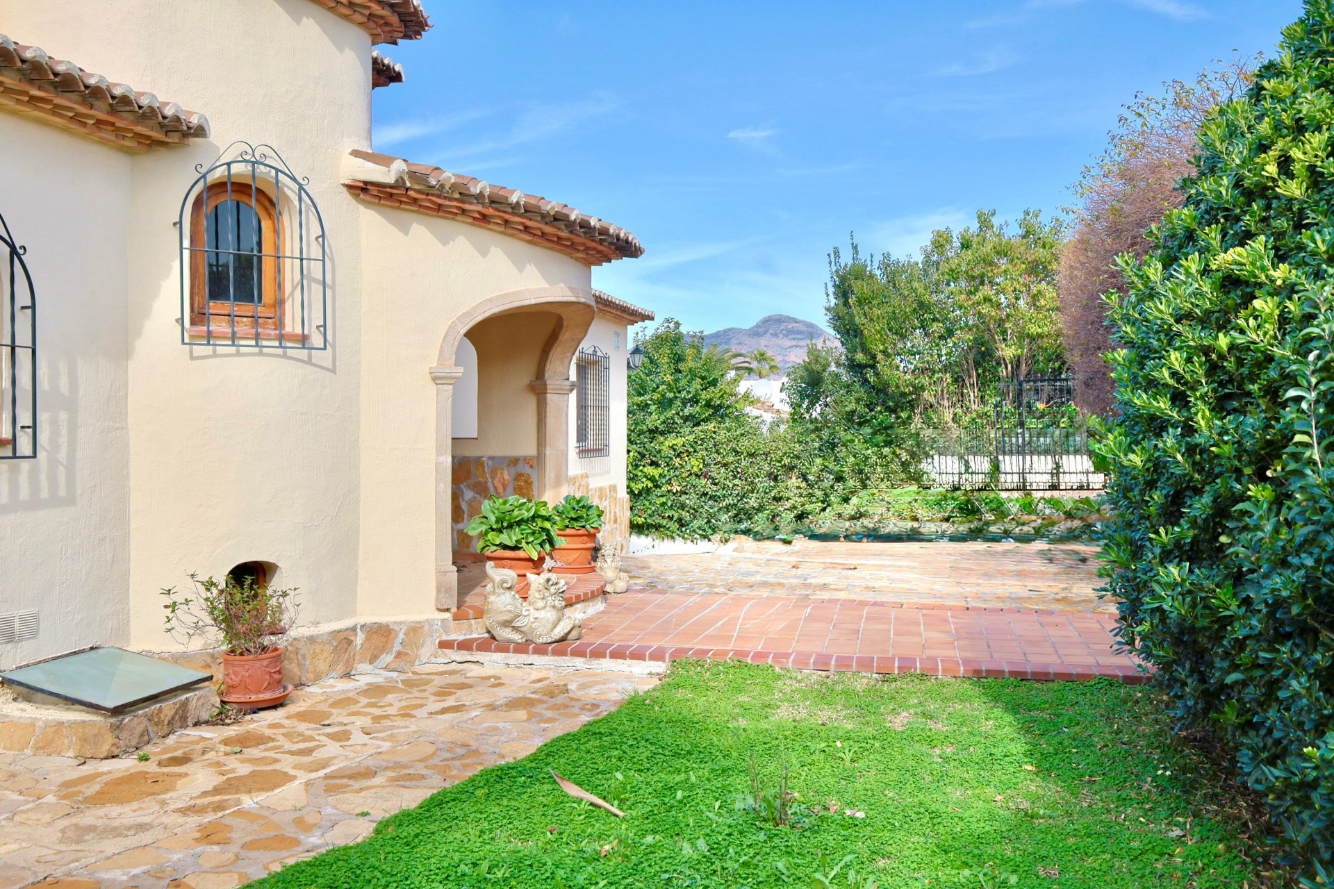 Exterior view of a Mediterranean villa featuring a stone-arched porch, terracotta tiling, and traditional wrought iron window grilles against a mountain backdrop.
