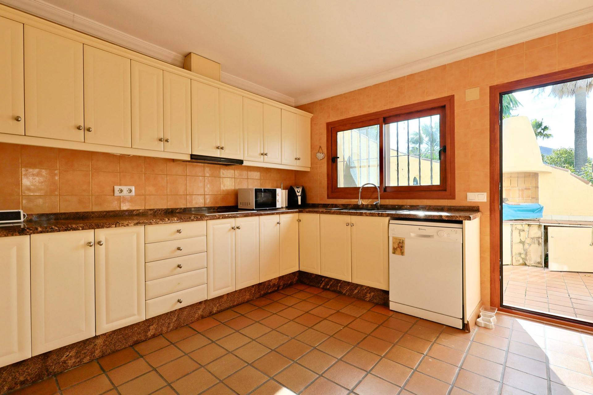 Fully equipped kitchen with cream cabinetry, granite countertops, and terracotta flooring. Features direct access to the terrace and a wooden-framed window.
