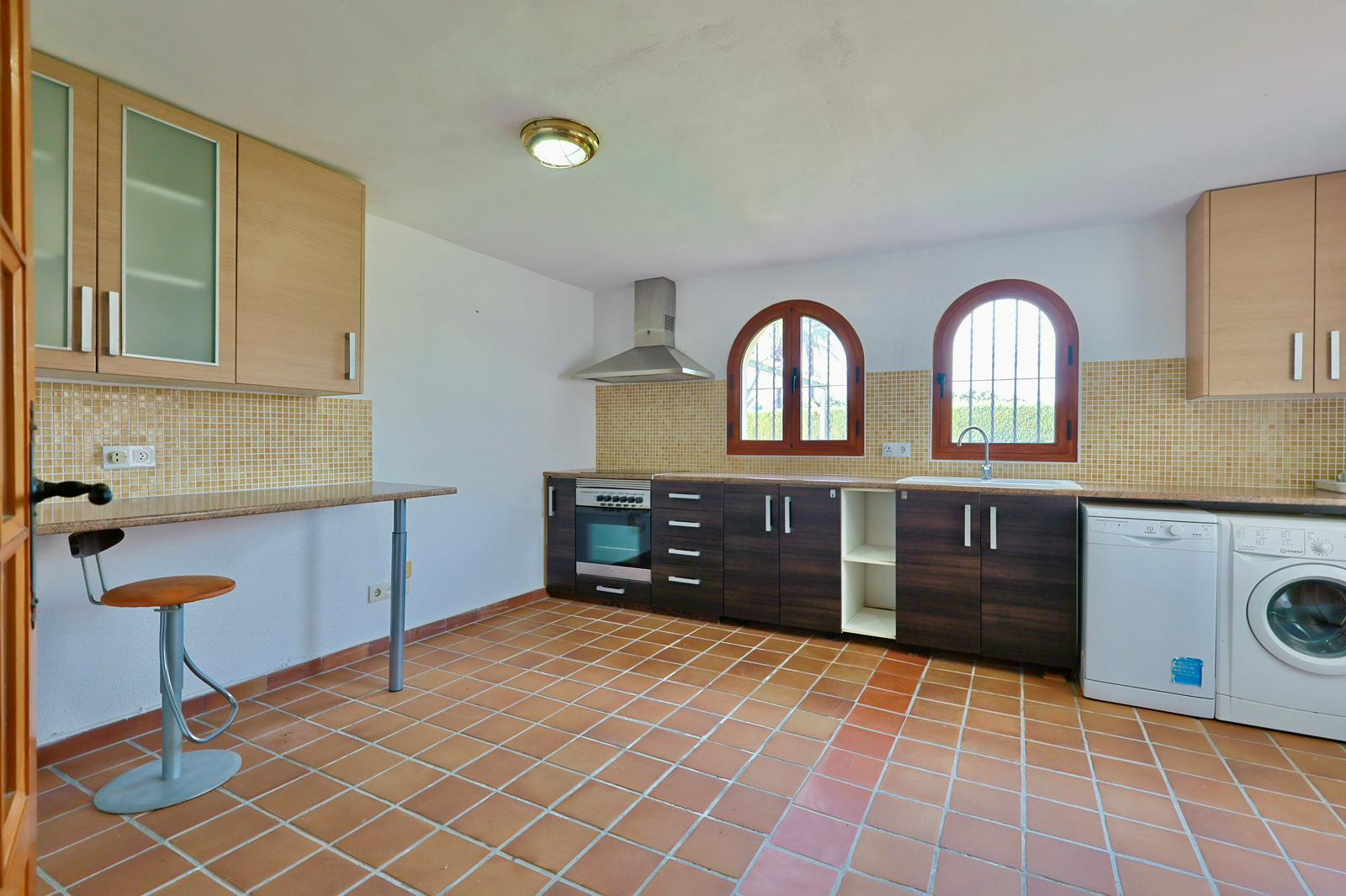 Functional kitchen featuring terracotta tile flooring, two-tone cabinetry, arched wooden windows, and integrated appliances. Includes a breakfast bar and beige mosaic backsplash.
