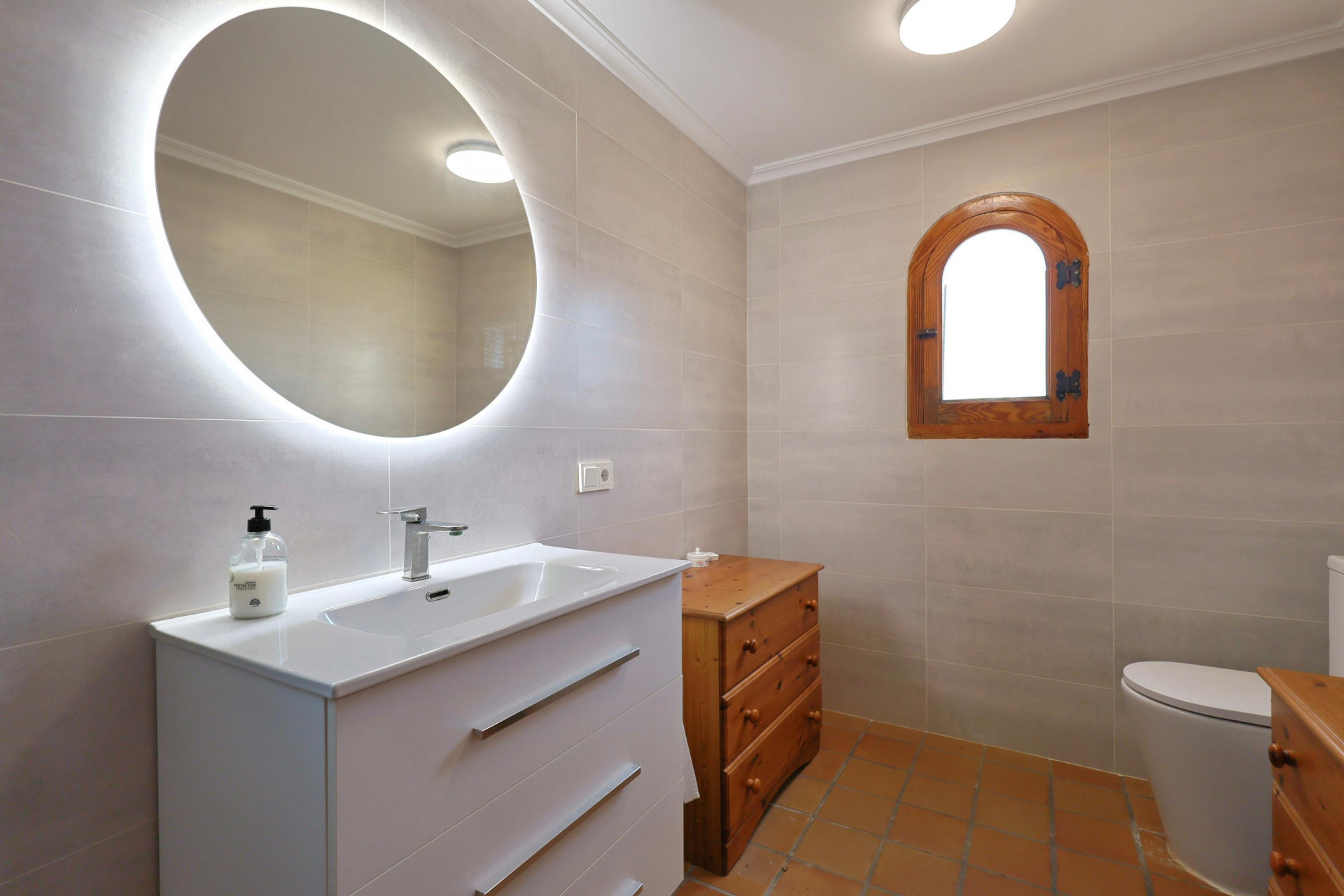 Modern bathroom featuring a backlit circular mirror, white vanity unit, terracotta tile flooring, and a traditional arched wooden window.