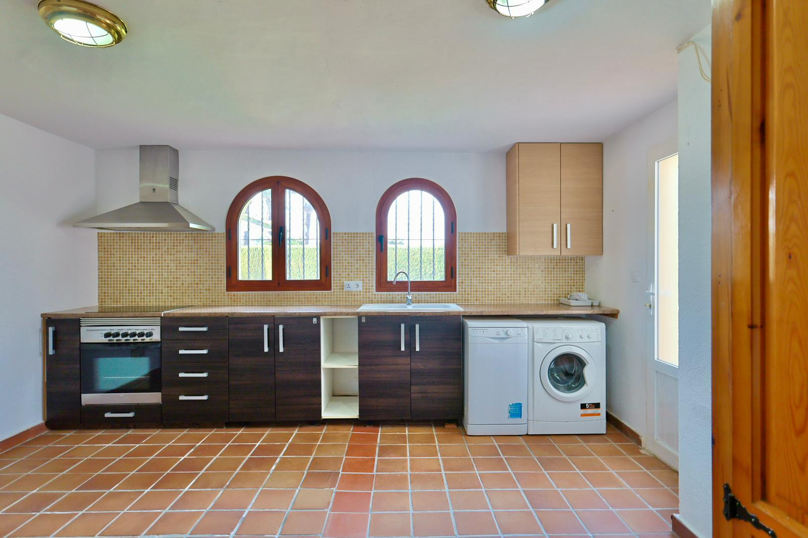 Practical kitchen and laundry area featuring dark cabinetry, terracotta flooring, and arched windows. Equipped with modern appliances and a mosaic backsplash.
