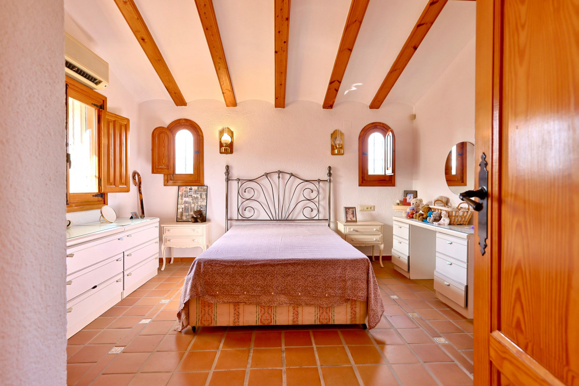 Rustic bedroom featuring exposed wooden ceiling beams, terracotta tile flooring, and arched windows with wooden shutters. Equipped with white cabinetry and a wrought iron headboard.