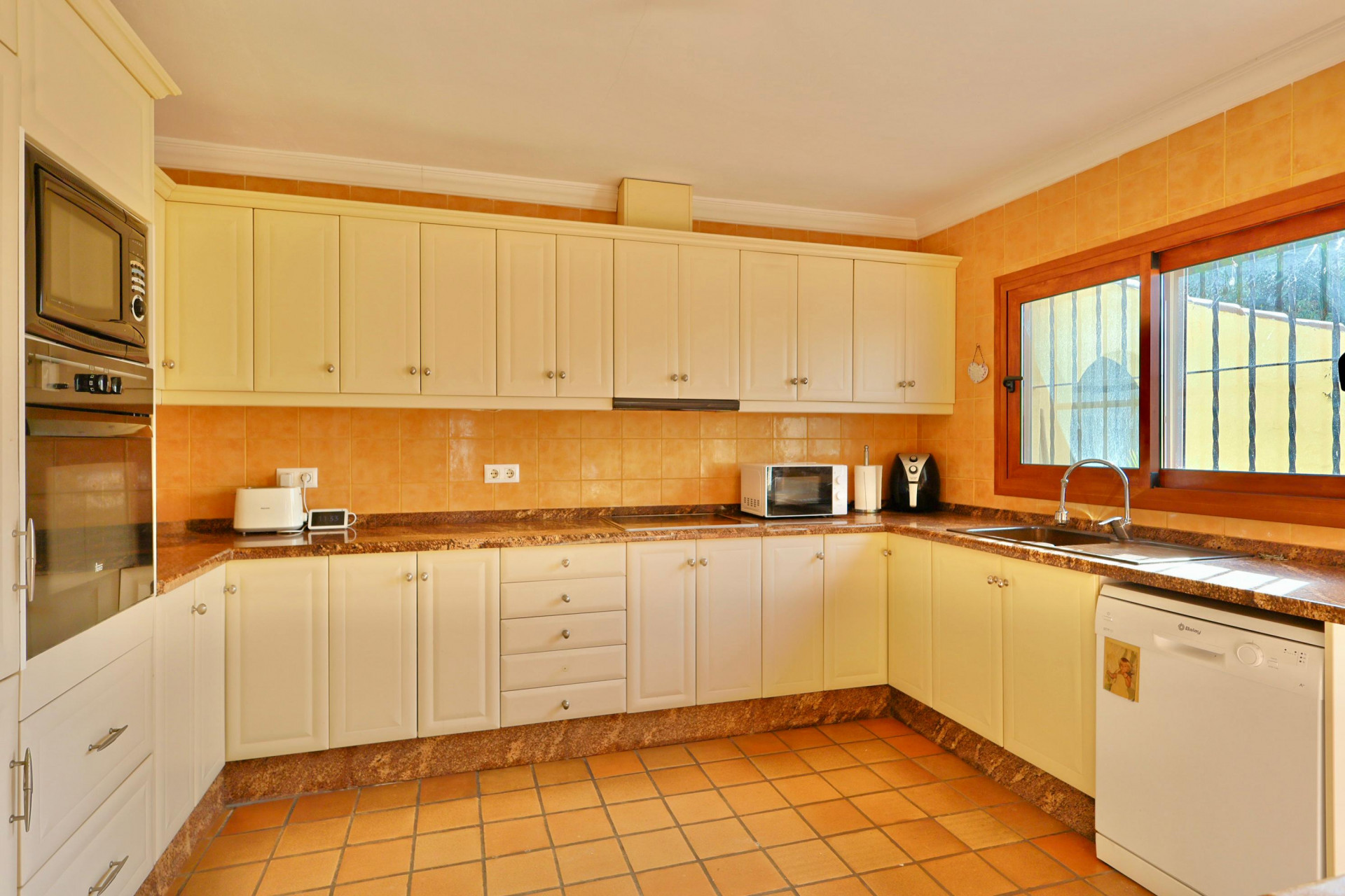 Traditional kitchen featuring cream cabinetry, brown granite countertops, and terracotta tiling. Includes integrated appliances and a wood-framed window.