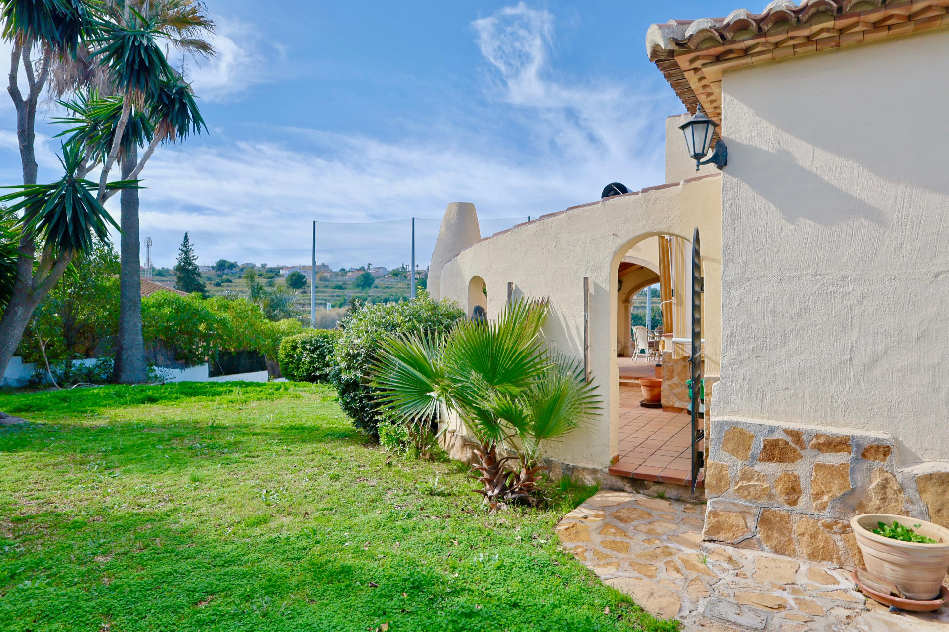 Villa exterior featuring a natural stone base, Mediterranean garden with palms, and an arched entrance to a terracotta terrace overlooking the golf area.