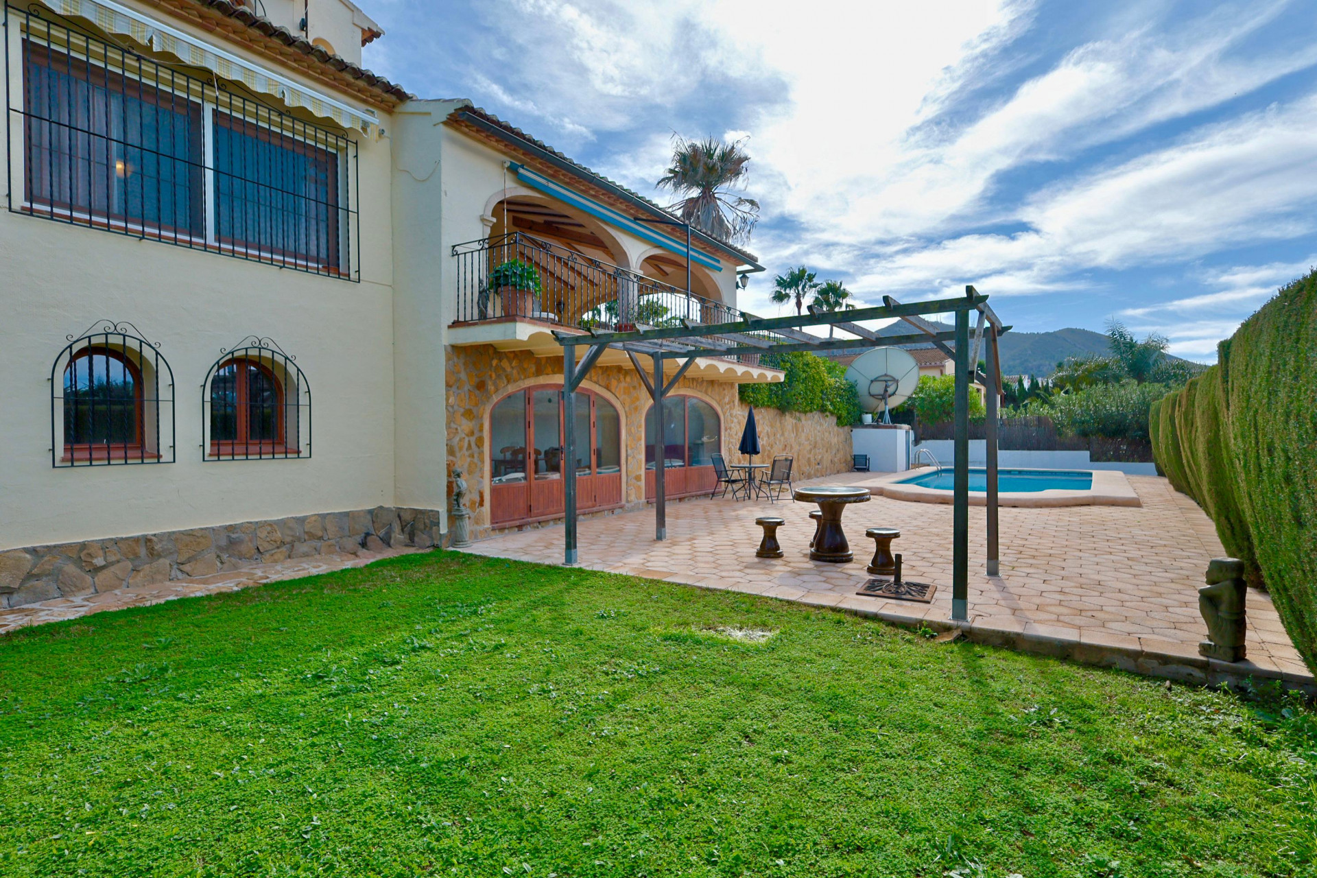 Villa exterior featuring a stone-clad lower level with arched doors, a wooden pergola on a tiled terrace, and a private pool beside a manicured lawn.