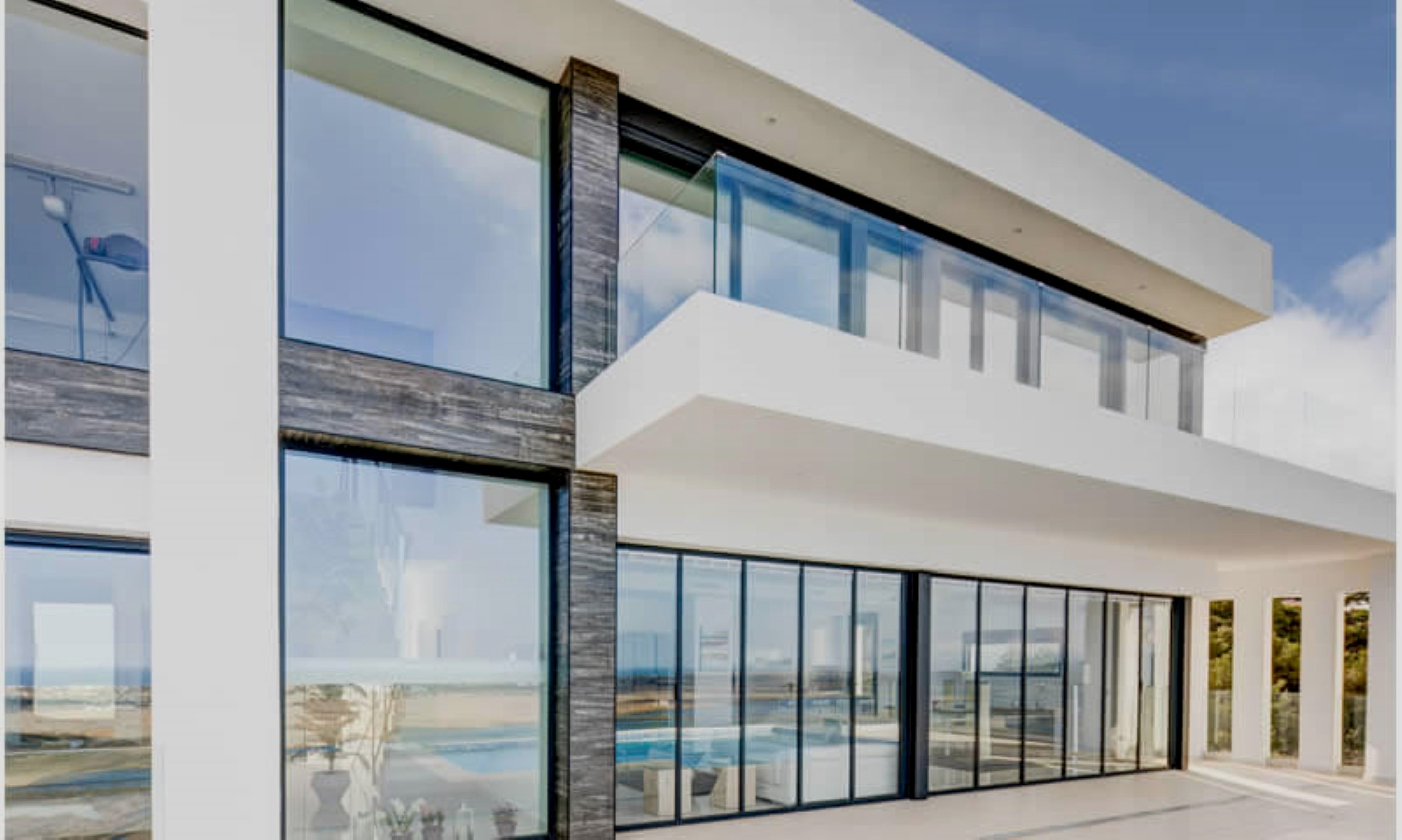 Modern facade featuring floor-to-ceiling glass panels, dark textured stone accents, and an upper-level balcony with a sleek glass balustrade.