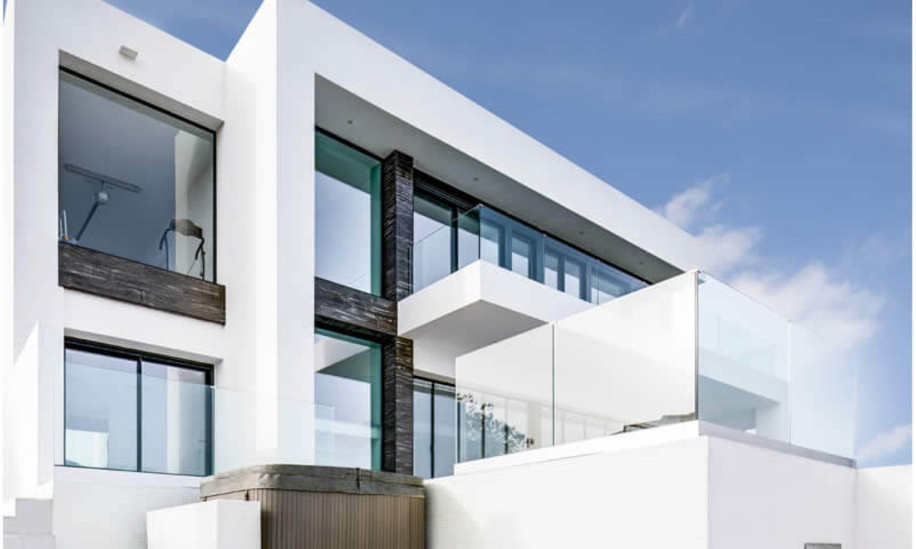 Modern geometric facade featuring white walls, large floor-to-ceiling windows, and glass balconies. Dark textured accents complement the sleek design and outdoor jacuzzi area.