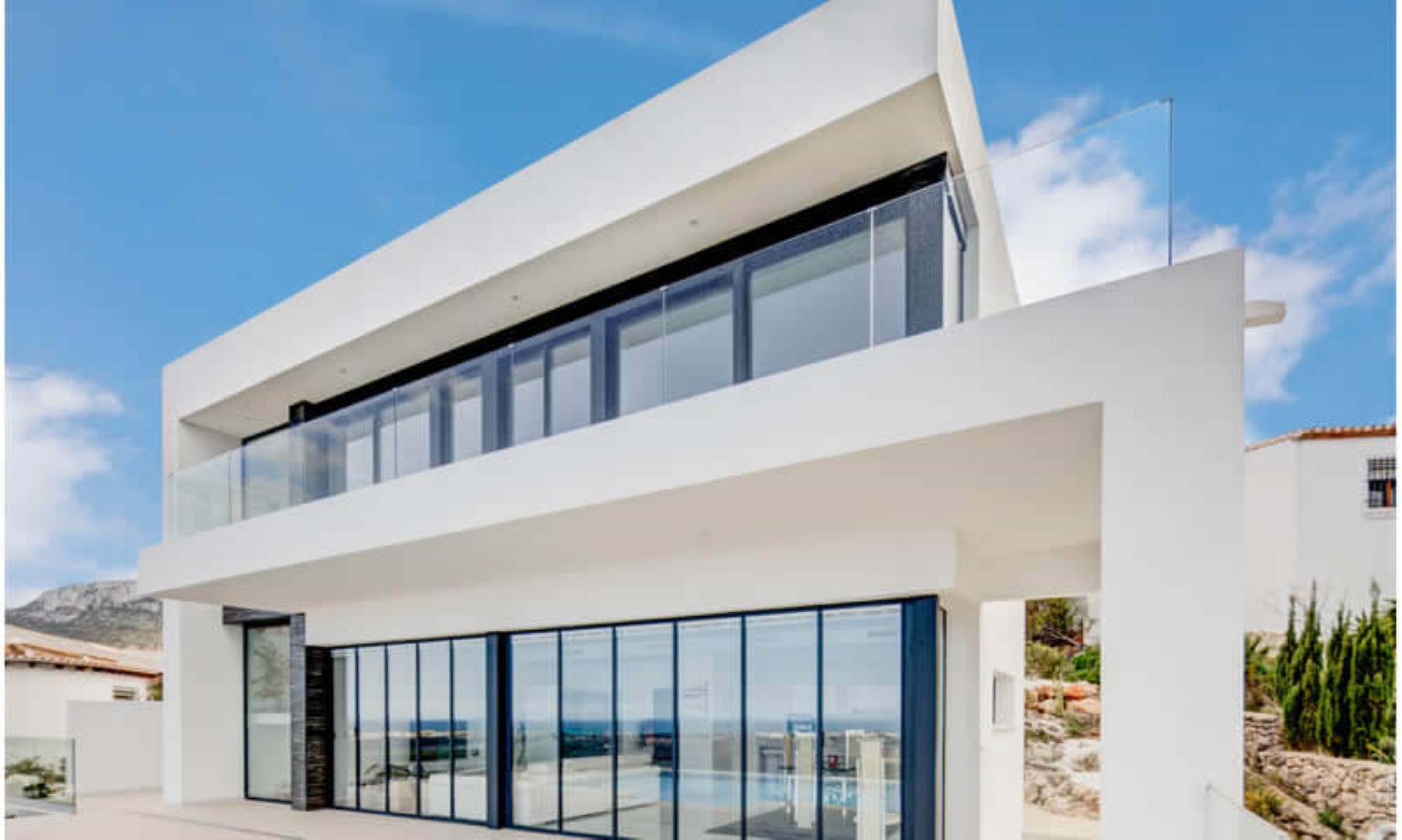 Modern two-story villa exterior featuring floor-to-ceiling glass walls, dark window frames, and upper-level balconies with frameless glass railings.