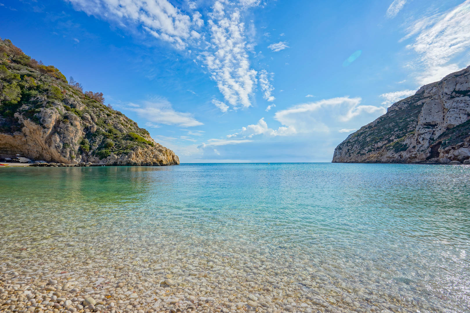 Panoramic view of Granadella Beach in Jávea, featuring crystal-clear turquoise waters, a white pebble shoreline, and surrounding rocky cliffs with Mediterranean flora.