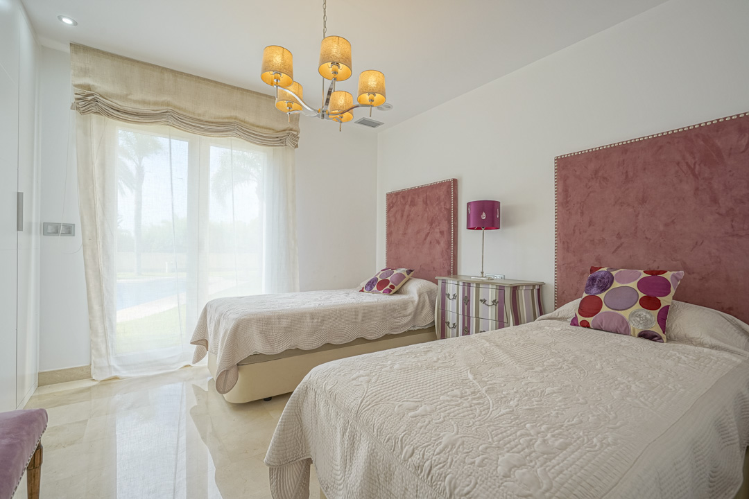 Bedroom with twin beds featuring pink velvet headboards and direct terrace access. Includes polished marble flooring, recessed lighting, and a classic fabric-shaded chandelier.