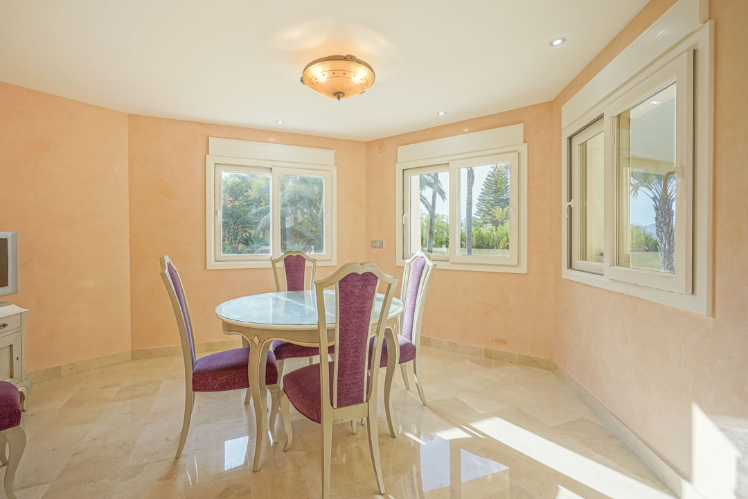 Bright dining area featuring a glass-topped table, purple upholstered chairs, polished marble flooring, and views of the Mediterranean garden through sliding windows.