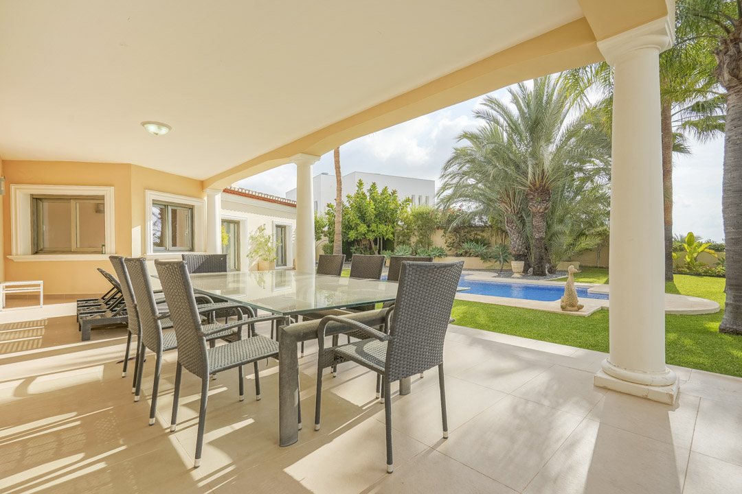 Covered terrace featuring white columns, a glass-top dining set, and views over the swimming pool and mature Mediterranean garden.