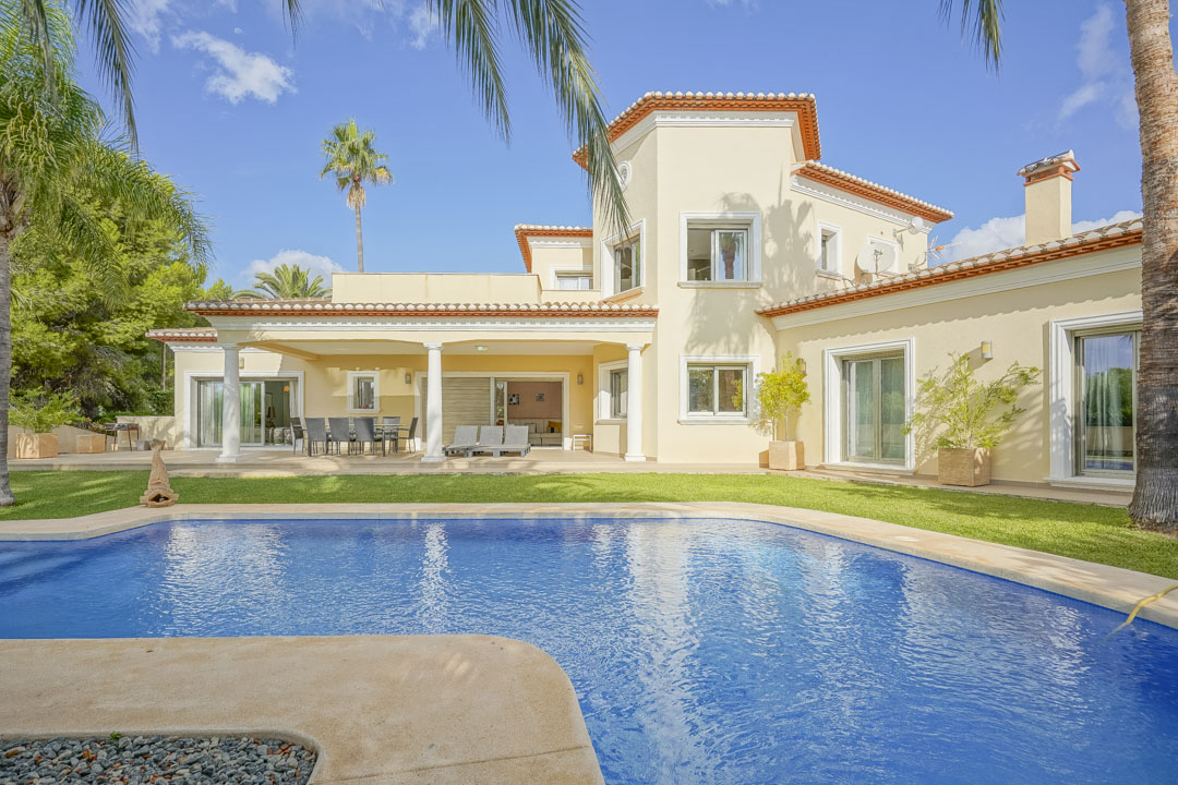 Exterior view featuring a large tiled swimming pool, a covered terrace with white columns, and a landscaped garden with mature palm trees.