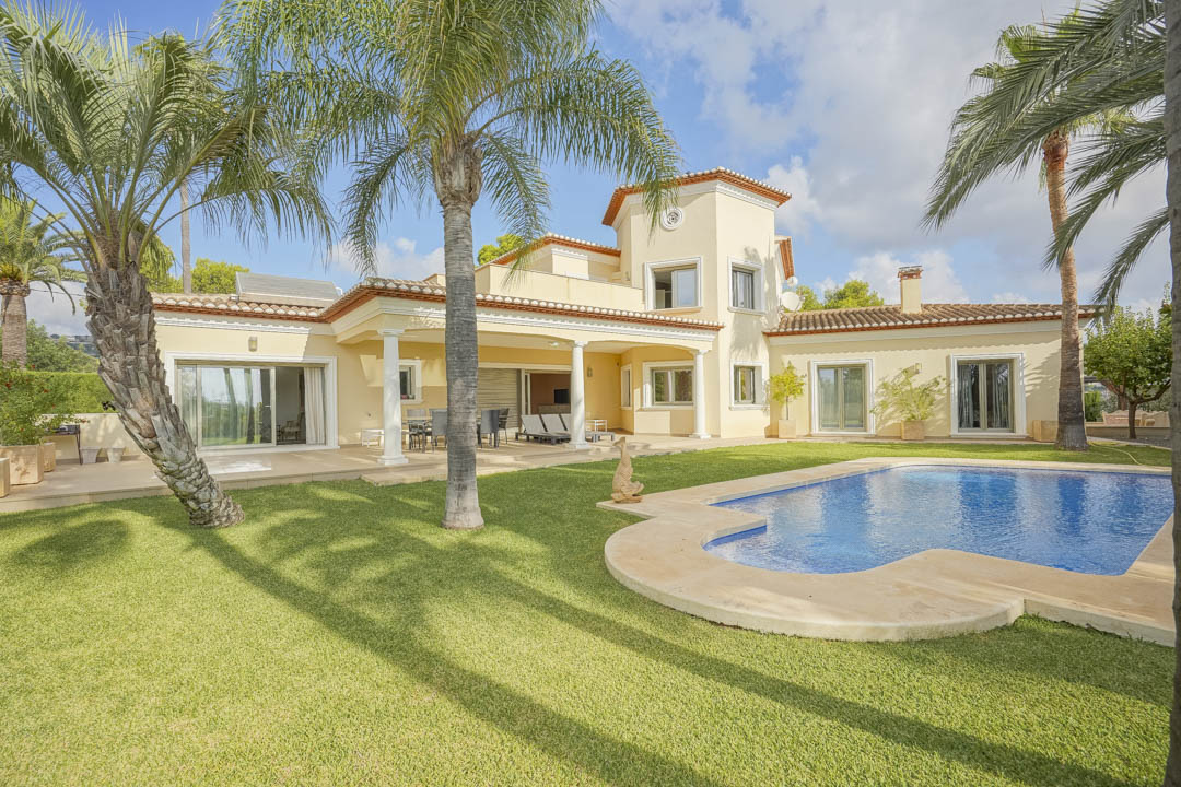Exterior view of the villa featuring a curved swimming pool, manicured lawn, and mature palm trees. The covered terrace with white columns connects the garden to the main house.