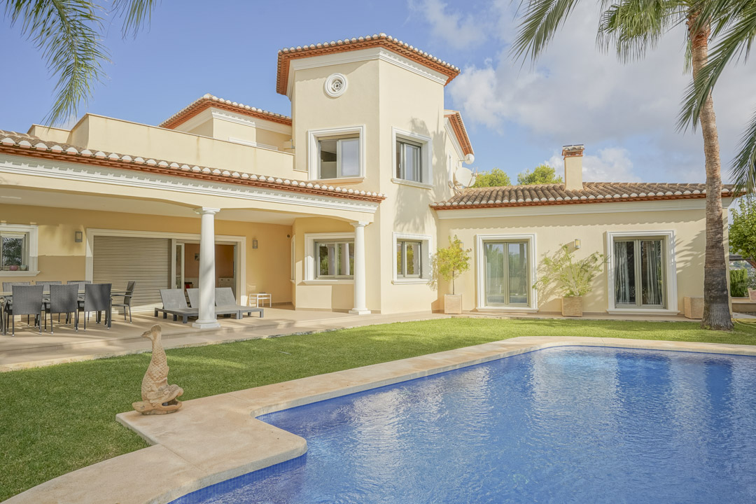 Exterior view of the villa featuring a large swimming pool, manicured lawn, and a covered terrace with columns and outdoor dining area.