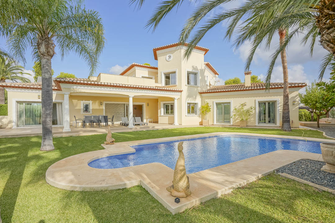 Exterior view of the villa featuring a large swimming pool, manicured lawn with mature palms, and a covered terrace with white columns and traditional tiling.