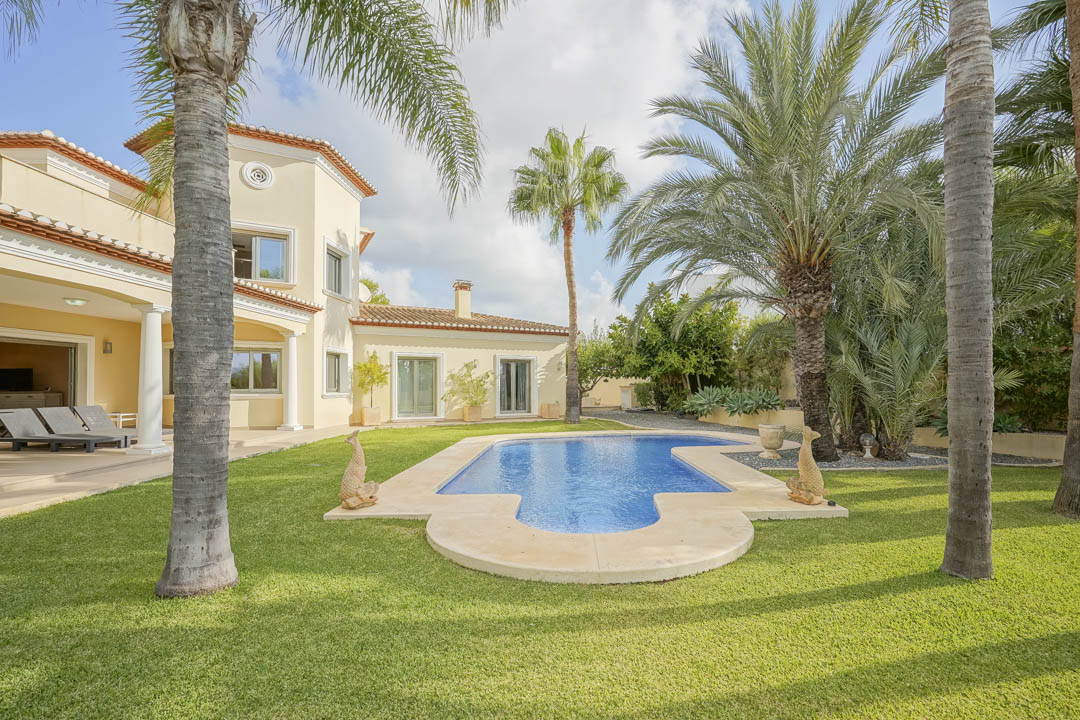 Irregularly shaped swimming pool set in a manicured lawn with mature palm trees, adjacent to a Mediterranean villa with a covered porch and white columns.