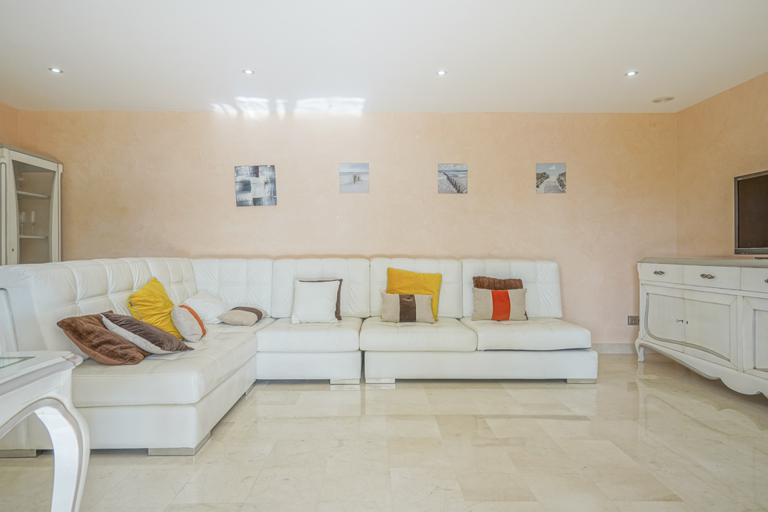 Living area featuring a white leather L-shaped sofa, polished marble flooring, and peach-toned walls accented by recessed ceiling spotlights.