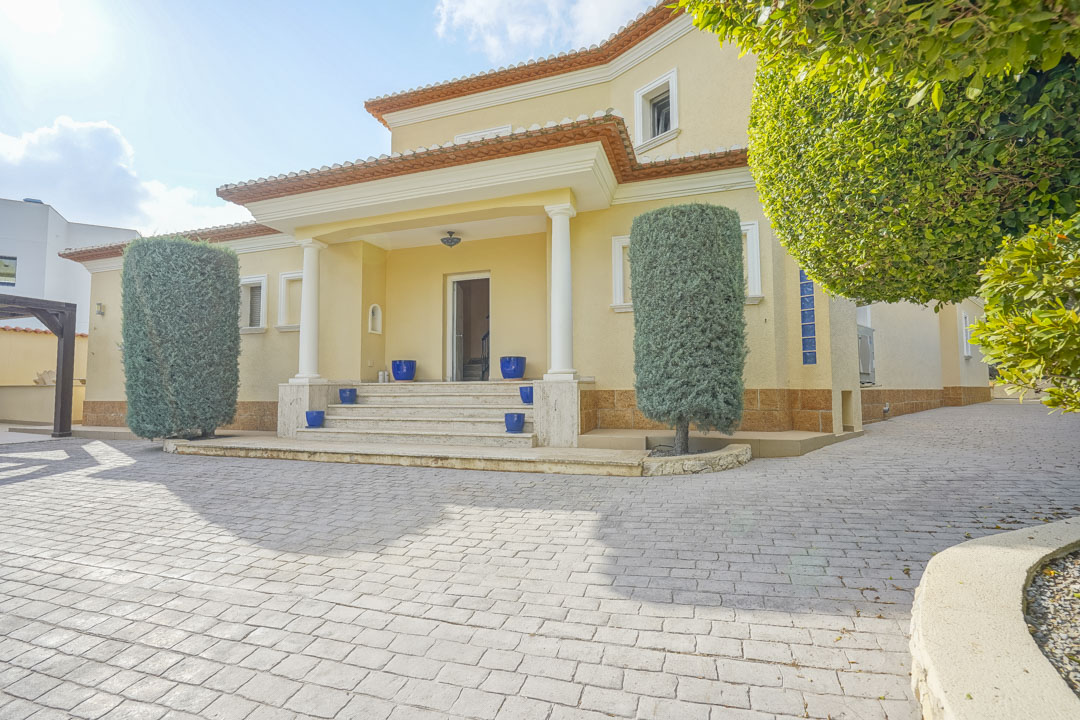 Main entrance featuring a columned porch, stone steps with blue ceramic accents, and a spacious paved driveway framed by manicured Mediterranean hedges.