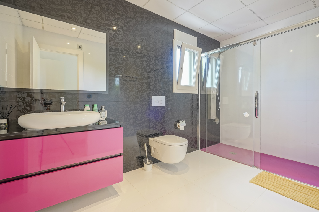 Modern bathroom featuring a vibrant pink floating vanity with a white vessel sink. It includes a walk-in glass shower with a matching pink floor and dark patterned accent wall tiles.