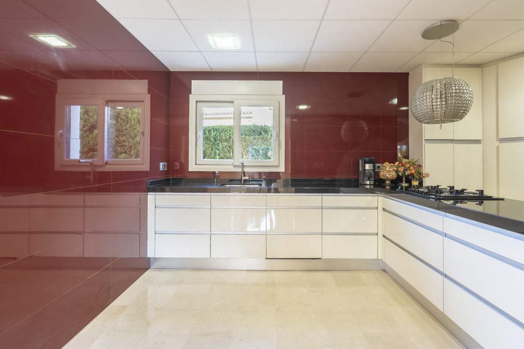 Modern kitchen featuring white handleless cabinetry, black granite countertops, and a bold red tiled accent wall. Equipped with a gas cooktop and a crystal pendant light.