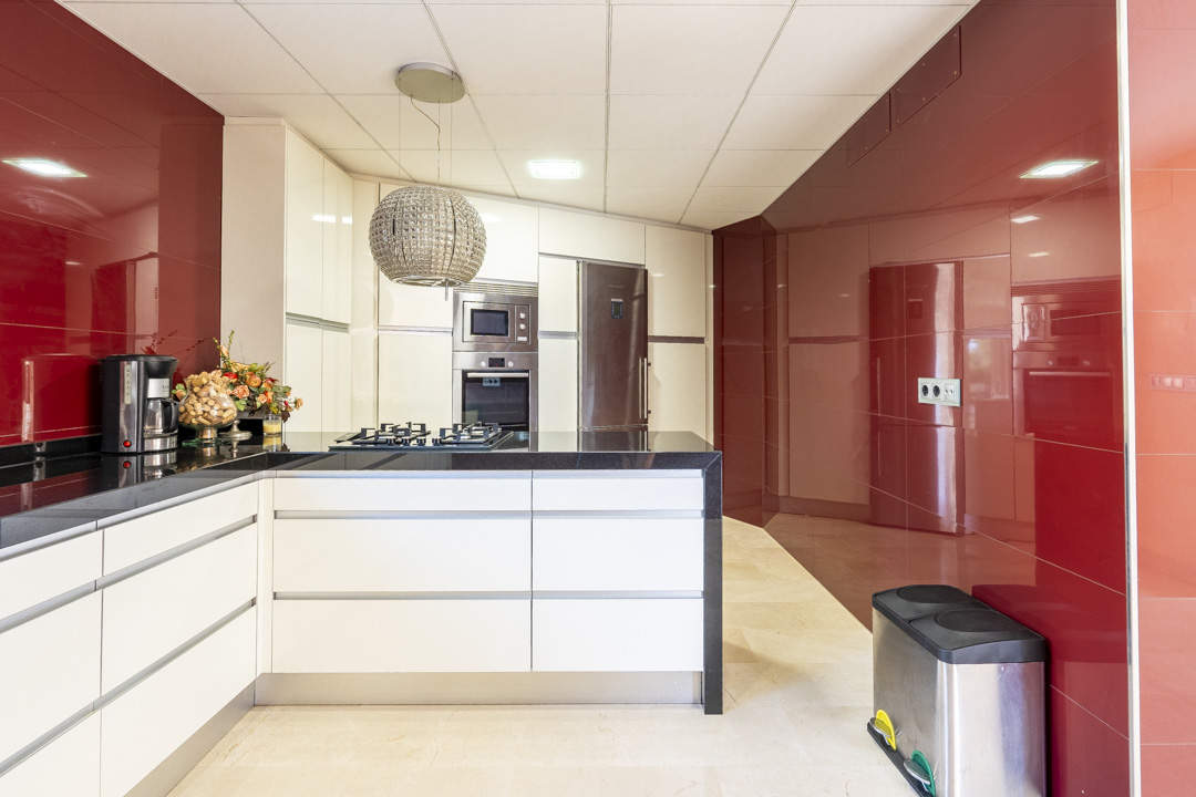Modern kitchen featuring white handleless cabinetry, black granite countertops, and glossy red wall panels. Equipped with integrated appliances and a decorative spherical pendant light.