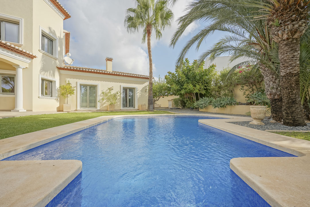 Outdoor swimming pool featuring stone coping, bordered by a manicured lawn and mature palm trees against the villa's cream facade and traditional tiled roof.