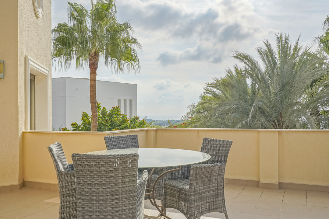 Outdoor terrace featuring a glass-top dining table and grey wicker chairs, framed by mature palm trees and distant sea views.