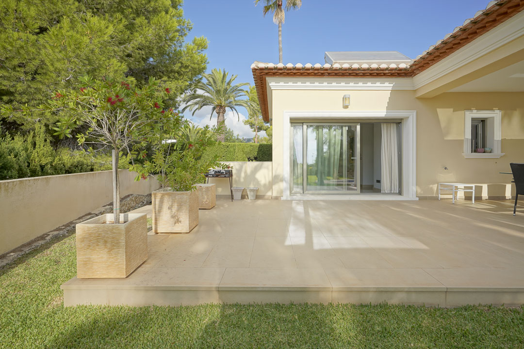 Outdoor terrace with light-toned tiling, direct sliding door access, and a garden featuring mature pines, palms, and rooftop solar panels.