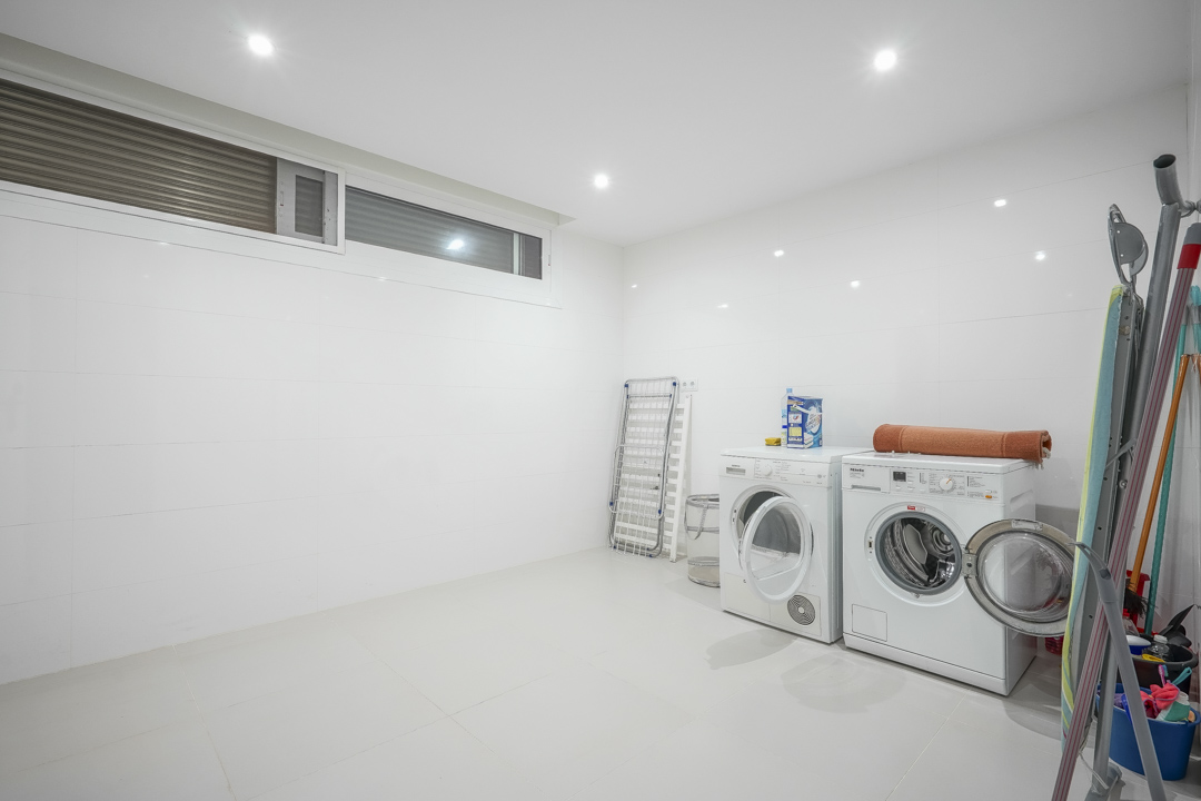 Spacious basement laundry room featuring white tiled walls, recessed lighting, and washing and drying appliances. Includes space for ironing and natural ventilation.