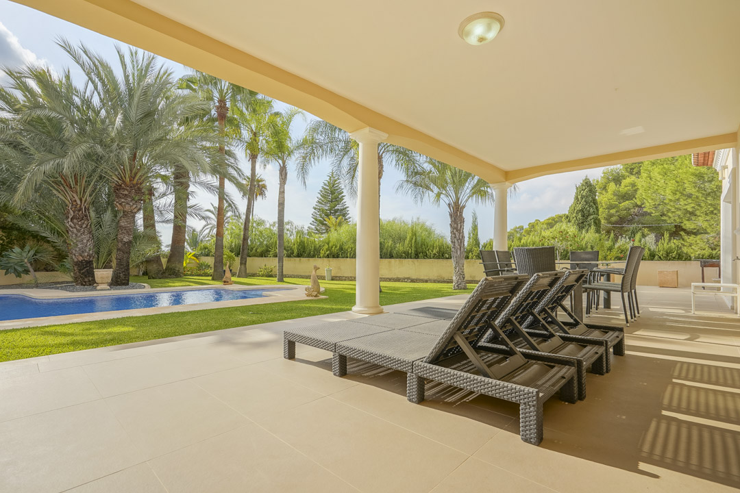 Spacious covered terrace featuring classical columns and outdoor furniture, offering views of the swimming pool and mature Mediterranean palm garden.