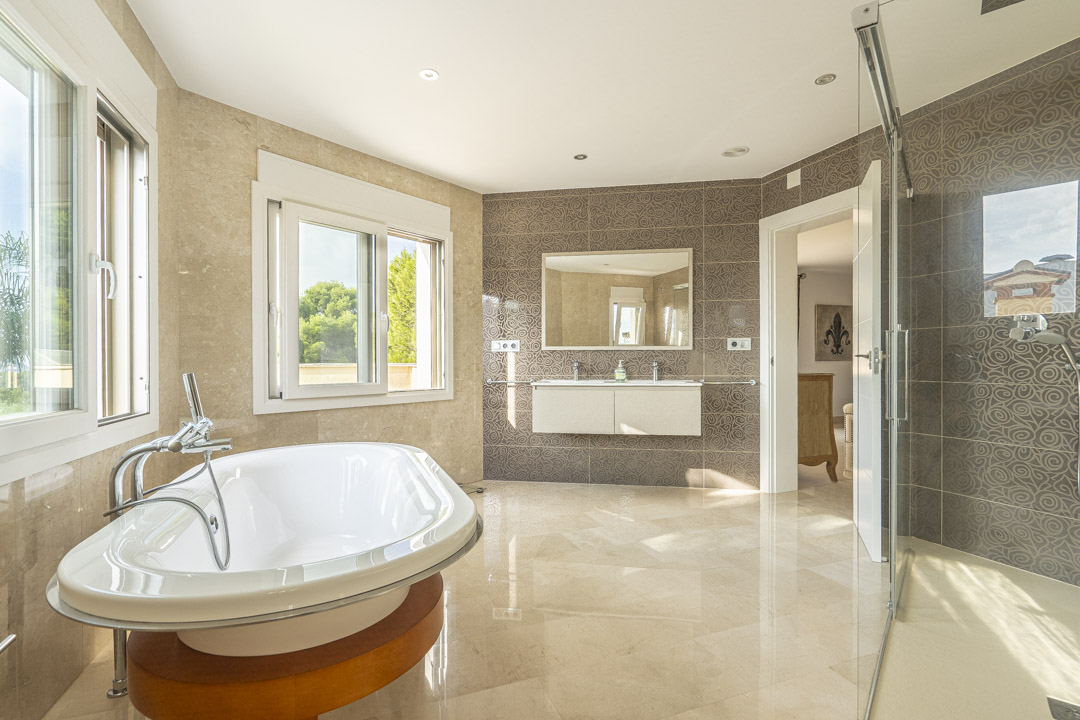 Spacious master bathroom featuring a freestanding oval bathtub on a wooden base, double vanity, and walk-in glass shower. Finished with marble flooring and patterned wall tiles.