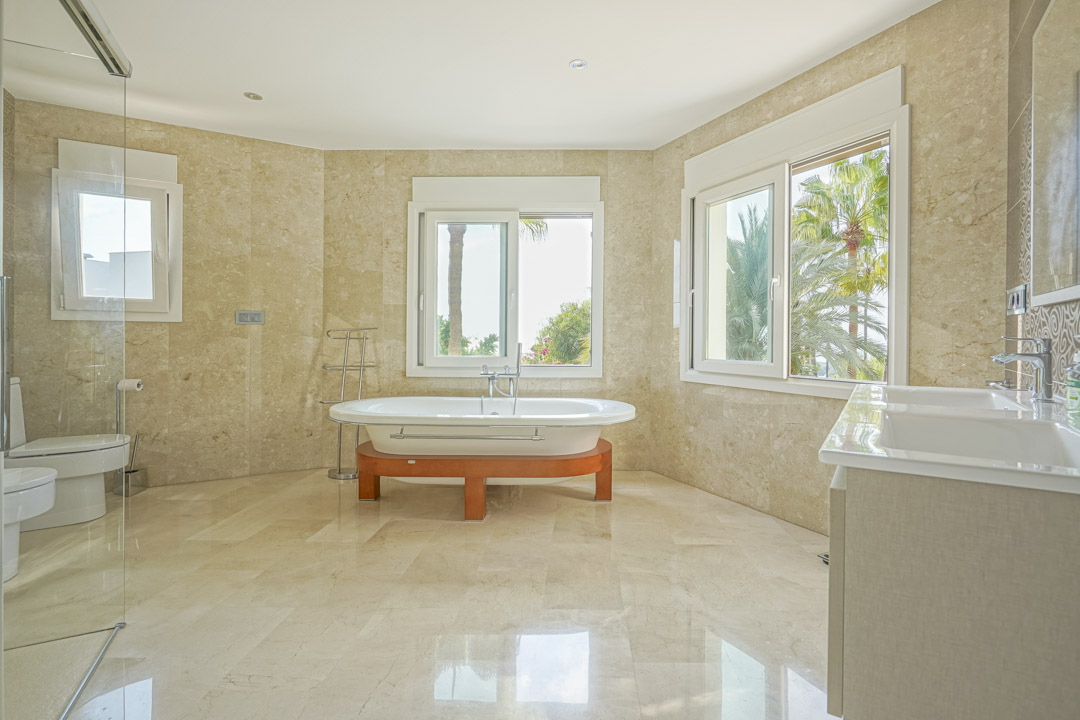 Spacious master bathroom featuring marble wall tiling, a freestanding oval bathtub on a wooden frame, and large windows overlooking Mediterranean palms.