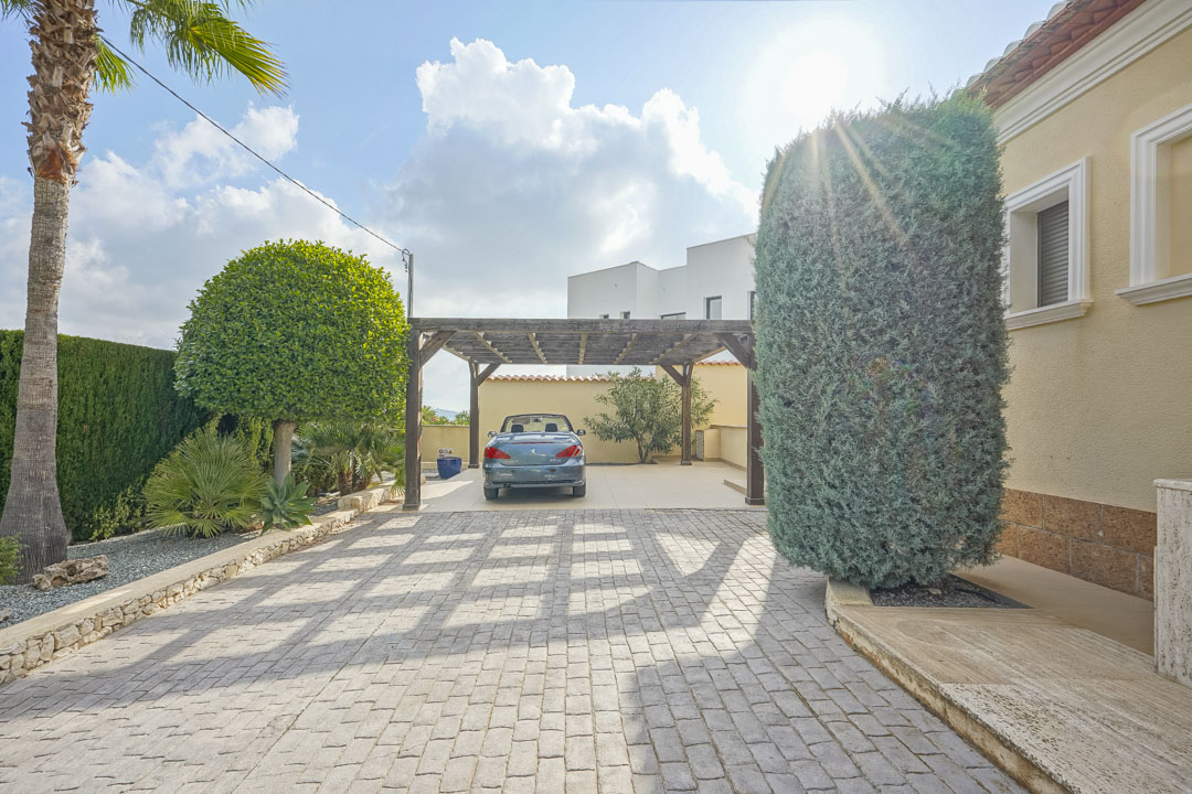 Spacious paved driveway featuring a wooden pergola carport, bordered by mature Mediterranean landscaping and a light-colored villa facade.
