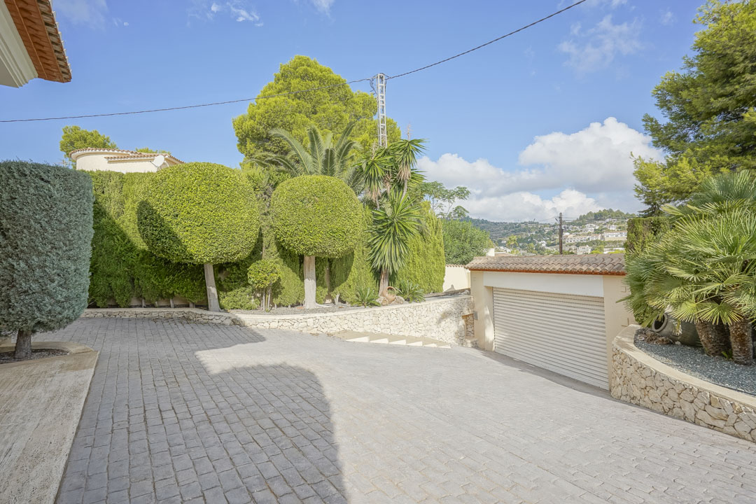 Spacious paved driveway leading to a garage, bordered by mature Mediterranean landscaping, topiary trees, and traditional stone retaining walls.