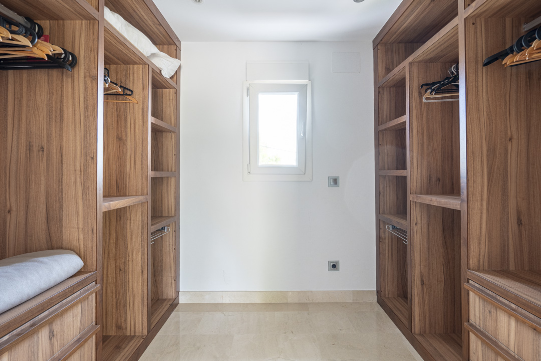 Walk-in closet featuring custom walnut wood cabinetry with open shelving and hanging rails. A central window provides natural light over the stone flooring.