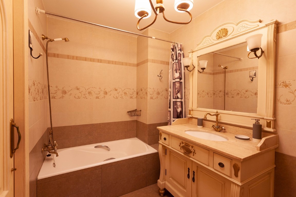 Classic bathroom featuring an ornate wooden vanity with a stone top and bronze fixtures. It includes a built-in bathtub and decorative cream-toned tiling.