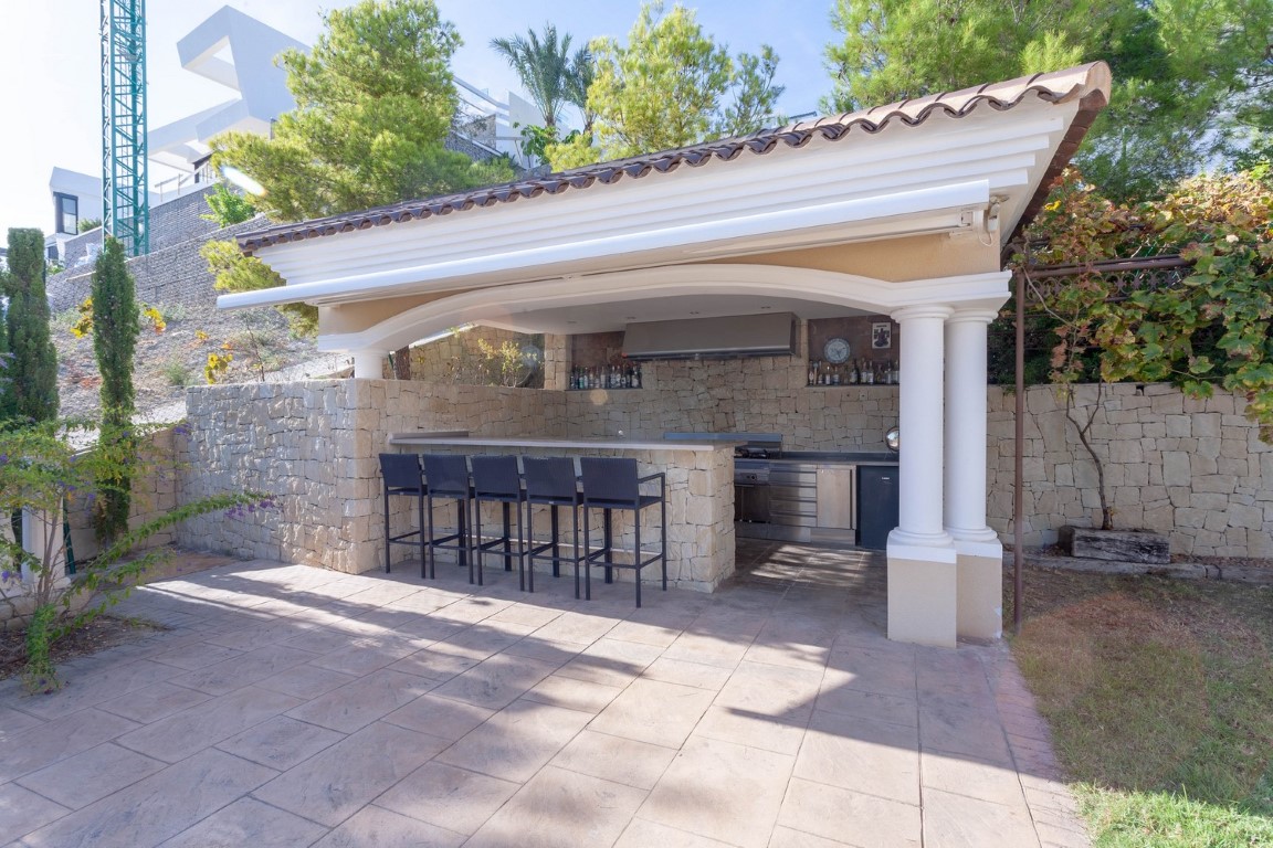 Covered outdoor kitchen featuring a stone breakfast bar with black stools, stainless steel appliances, and traditional tiled roofing supported by columns.