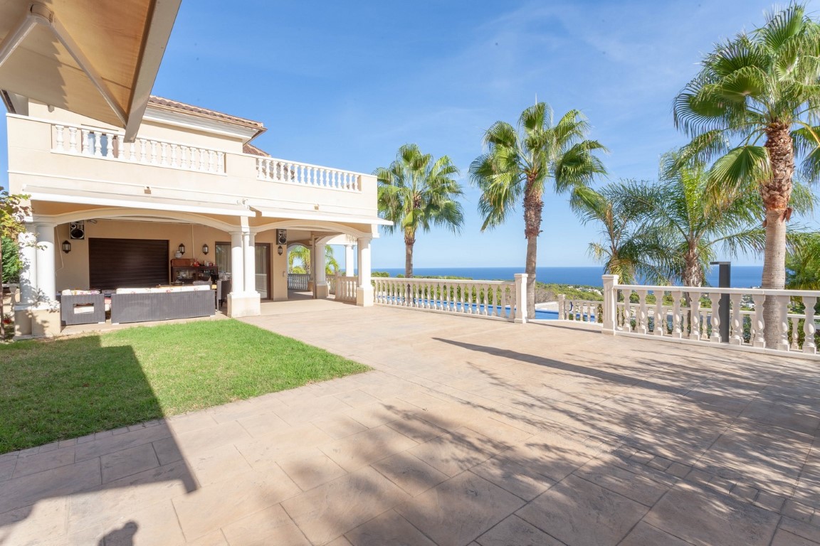 Expansive stone terrace featuring classic white balustrades and panoramic Mediterranean views. The space includes a covered porch with seating and mature palm trees.