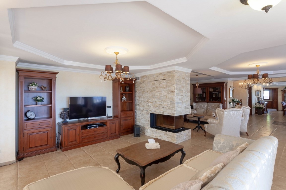Ground floor living room featuring two separate seating areas, a central stone-clad fireplace, classic dark wood cabinetry, and tiled flooring.